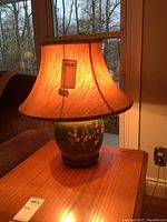 Lamp fully lit showing illumination through silk shade and ceramic base on wooden table near window.