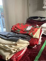 Assorted table linens stacked, featuring red Christmas print, cream textured fabric, and dark gray vinyl or fabric.