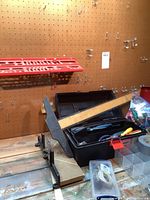 Two black toolboxes with one open showing a yellow screwdriver inside, red wall-mounted tool holder, and wooden ruler clamped to the table.