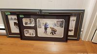All three framed prints leaning against a wall on a wood floor. Two larger prints show snow scenes with children and trees, one smaller print frame shows three smaller images with children playing in snow.