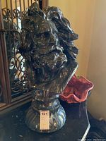 Front view of dark greenish-black ceramic bust with long hair and beard, mounted on pedestal, showing size and shape.