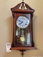 Full front view of the London regulator style wall clock showing wooden case, dial with Roman numerals, and pendulum behind glass door.