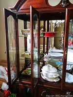 Wide view of wooden china cabinet with glass doors displaying white soup tureen, matching bowls and plates, red cake stand, and glass jam jars.