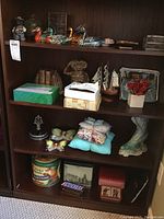 Shelf showing various miniatures and figurines including wooden ducks, a ceramic or resin dog figurine dressed as a cowboy, decorative tins, pillows, bookends, a model ship, a glass candy dish, and other small decorative items.