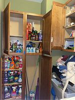 Wide view of wooden cabinets with shelves containing assorted cleaning bottles, sprays, folded linens, and cleaning tools like mops and brushes.