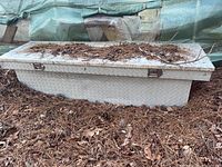 Metal toolbox with diamond plate pattern, viewed from front and side angles, partially covered with natural debris like pine needles and leaves. The toolbox is sitting outdoors on a bed of dried pine needles.