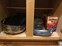 Photo of swivel cabinet shelf holding a large round roasting pan with rack, a metal oval baking pan, a cylindrical metal pot/lid, and a boxed Magic Brownie Pan.