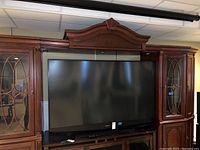 Front view of the Mitsubishi flat screen TV showing the screen and its stand placed on a wooden surface with a large wooden cabinet in the background.