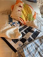 Four quilts and a flannel duvet displayed folded on a bed showing various patterns and colors including blue and white star design, black and white patchwork, and orange and cream with boat motifs.