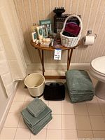 Photo shows four green bath towels, four hand towels, two washcloths, a white trash can, a black Health o meter scale, and a wooden side table with various bath products including an Infiniti Pro Conair hairdryer with diffuser.