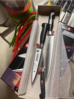 Henckels 3-piece knife set with black handles in protective packaging shown with colored tongs in background