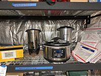 Kitchen appliances and accessories on metal shelving including crock pot, coffee maker, beverage dispenser, thermometer, and USPS Priority Mail envelopes.