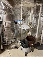 Photo of metal folding drying rack, two-tier rolling clothing rack with hangers, mop wash tub, and bucket with mop wringer in basement setting.