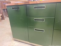 Photo of green metal office credenza section showing cabinet door with inset metal pull, two drawers above a larger bottom drawer, resting on a silver metal base.