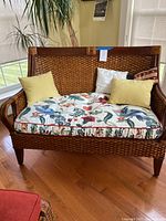 Wood and rattan loveseat with multicolored tropical floral and bird pattern seat cushion and four throw pillows (two yellow, two white including one with a red/beige pattern). Showcases woven rattan back and base with solid wood framing in medium brown finish.