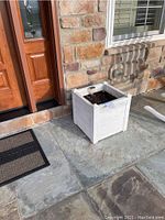 Single white square planter filled with soil on outdoor stone floor near front door.