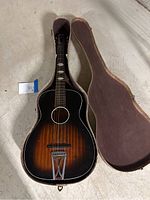 Acoustic guitar inside the opened hard case on the floor, showing sunburst finish and trapeze tailpiece.