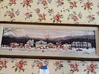 Long horizontal framed painting depicting a winter town scene with multiple buildings and figures, likely a Norman Rockwell style representation of Stockbridge, Massachusetts.