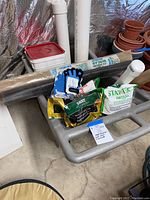 Full overview of the wheelbarrow showing the black garden fabric roll, gloves, fertilizer bags, and white container inside, situated in basement with pots nearby.