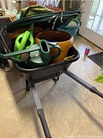 Wheelbarrow loaded with two clay-look plastic planters, two green watering cans, and green bush covers