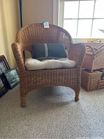 Full front view of wicker chair with gray pillow and white seat cushion on carpeted floor near window.