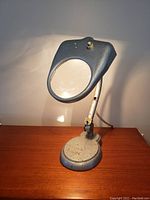 View of the lamp angled to the left, showing the lamp head, magnifying glass, and base on wooden surface.