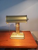 Front and side view of antique brass-colored desk lamp on wooden surface showing cylindrical shade and chain pull switch.