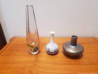 Lot of three vases on wooden surface: tall clear art glass vase, small white and blue porcelain vase, short round art glass vase.