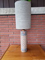 Full view of vintage tall ceramic table lamp on wood surface, showing detailed patterns on ceramic base and the tall gray fabric lamp shade.