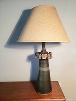 Full view of the lamp, showing conical ceramic base in varying brown and olive tones and beige woven lampshade.