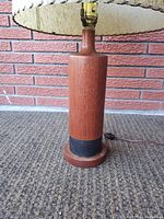 Full view of the lamp on carpet in front of a brick wall, showing teak wood base, black band near the bottom, and the top half of the cream textured fabric lampshade.