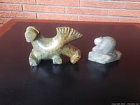 Photo showing front and side views of two soapstone animal carvings on black surface against brick background.