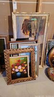Framed prints stacked on the floor showing a large print of a historical building with some damage to the frame and a print of a blue vase with multicolored flowers.