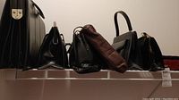Photo of various black bags including briefcase style bags, side bags, and wallet or portfolio bags lined up on shelf.