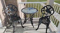 Photo showing two black metal bistro chairs with decorative floral design and a round metal table with a glass top on a porch.