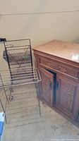 Wood cabinet with carved floral design doors and drawer, next to two black metal wire racks. The cabinet shows a flat top and dark metal handles.