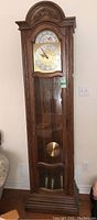 Full frontal view of the tall wooden grandfather clock showing carved arch, glass door, pendulum, and weights.