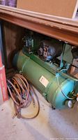 Photo showing the side view of the green air compressor tank connected to motor and pump with several coiled hoses.