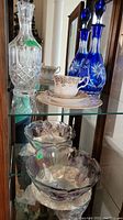 Glass shelf showing full lot including blue crystal decanters, clear crystal decanter, Royal Albert teacup and saucer, and glass bowls.