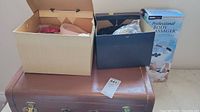 Lot overview showing two hat boxes on top of a brown suitcase with an unopened massager box behind