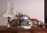 Full view of lot items on table including metal lamp with fabric shade, metal chandelier lamp, clock, decorative plates, flowers, and phone.