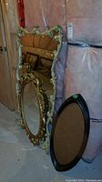 Two ornate mirrors with detailed gold and cream carved frames and one oval black wooden frame leaning against a wall in a basement space with insulation background.