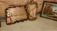 Two heavy ornate gold-tone framed mirrors leaning against a wall with one visible painting beside them.