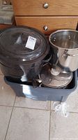 Photo showing large black enamel speckled pot in blue plastic bin with various cookware and a large stainless steel stock pot with lid beside it.