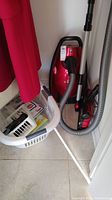 Red Samsung Ultra Quiet Jet canister vacuum cleaner standing on a tiled floor next to a white laundry basket containing various vacuum bags and accessories. Vacuum hose and attachments visible.