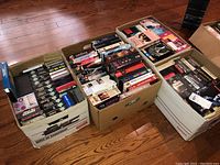 Four cardboard boxes filled with VHS tapes on a wooden floor, showing dense packing with various movies and series.