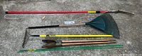 Full view of assorted landscaping tools including large rake, landscaping rake, post hole digger, limb trimmer pruner, and metal stakes.