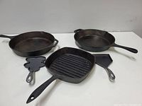Five cast iron skillets of various sizes and shapes arranged on a white surface under neutral lighting. Visible are two round pans, a square ridged grill pan, a small clover shaped mini skillet, and a small square pan.