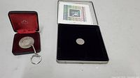 Two collectible items: a silver commemorative coin attached to a keychain in a red velvet box with a Canadian maple leaf symbol, and a commemorative 2000 coin stamp in a black velvet case with a clear cover.