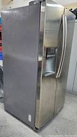 View of the front and left side of the stainless steel fridge, showing the external water and ice dispenser and vertical handles.